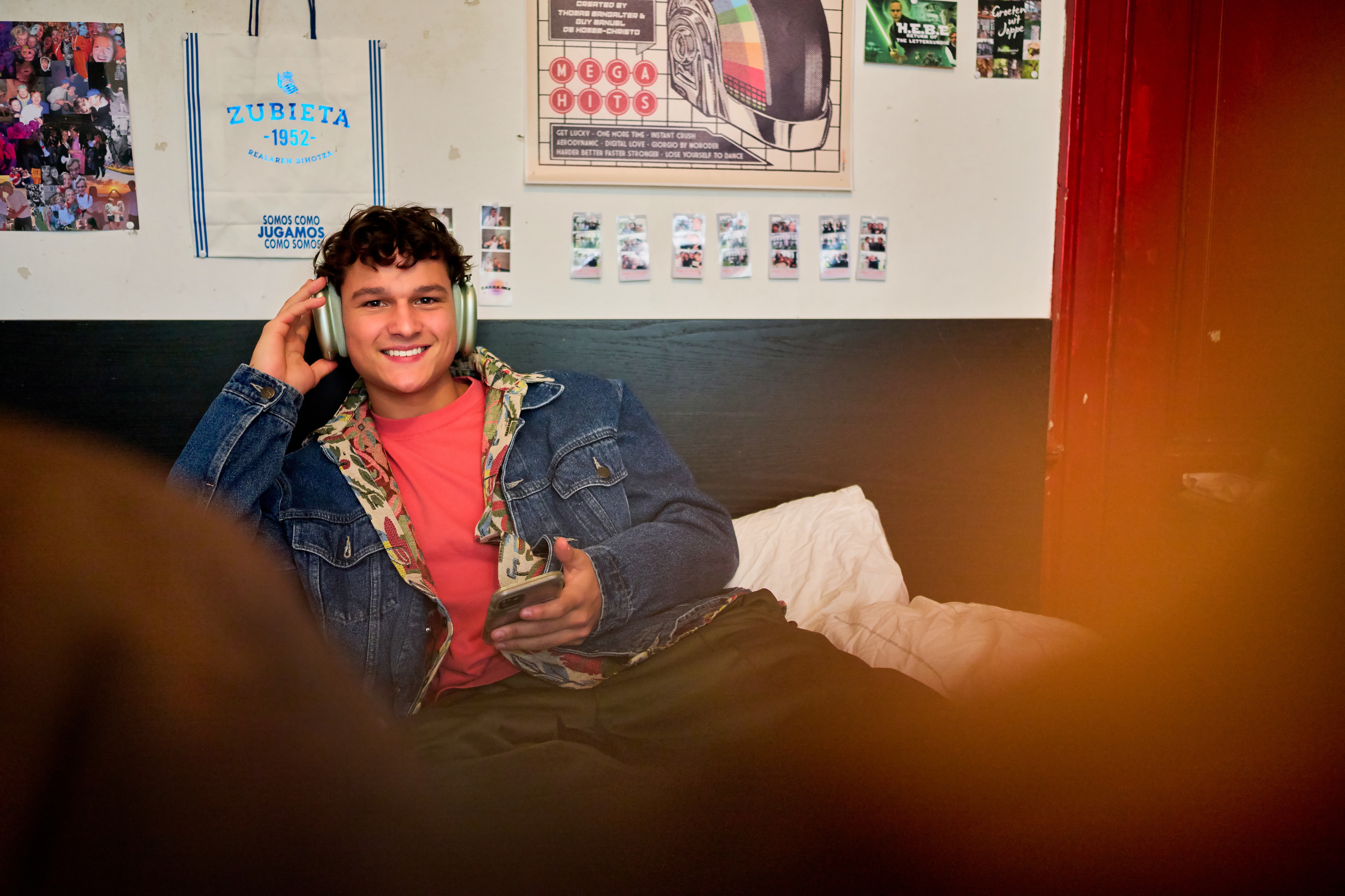 Boy is sitting on his bed with headphones on and phone in his hand, looking at the camera.