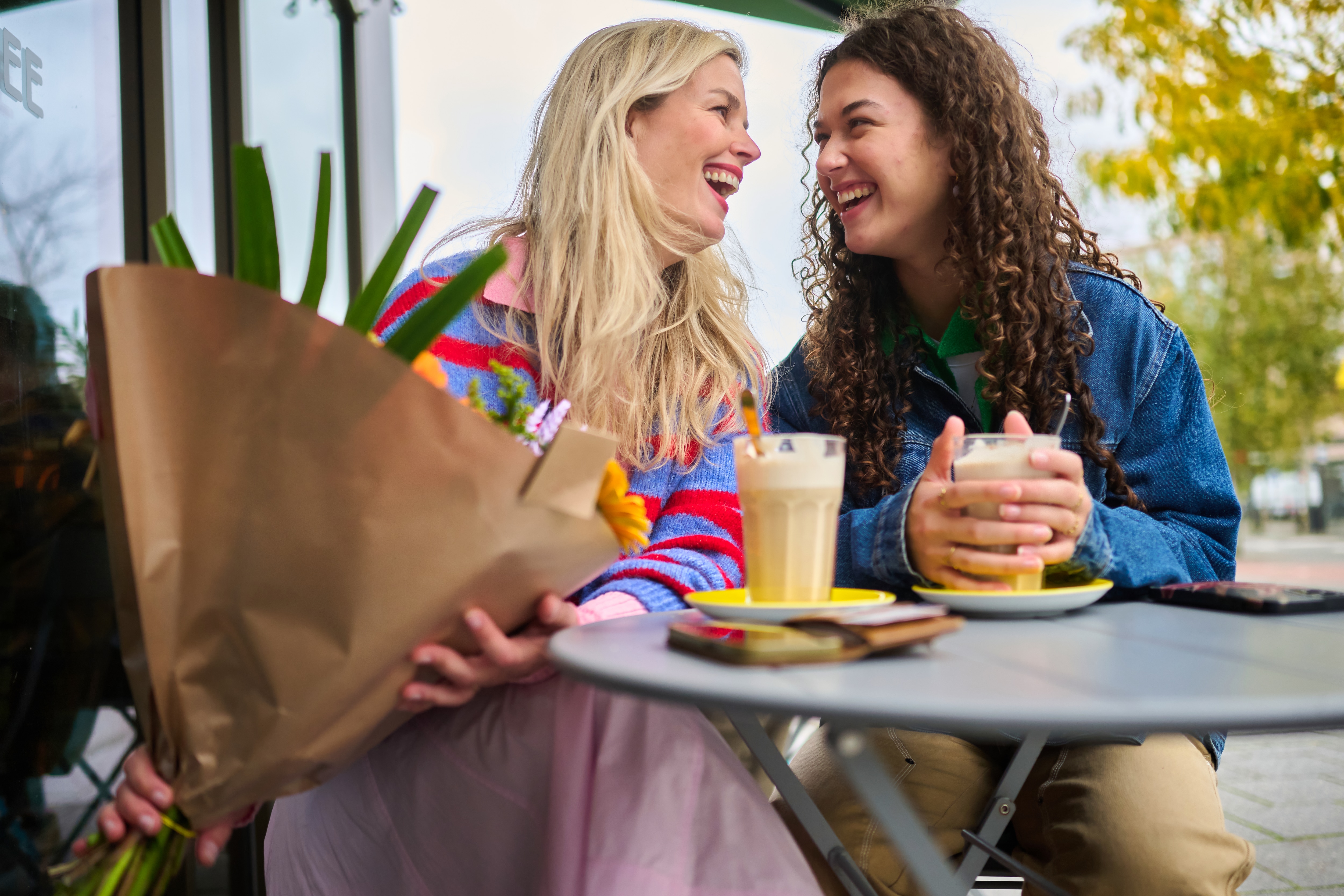 Two women sitting outside at a café table, talking and laughing together; one woman is holding a bouquet of flowers.