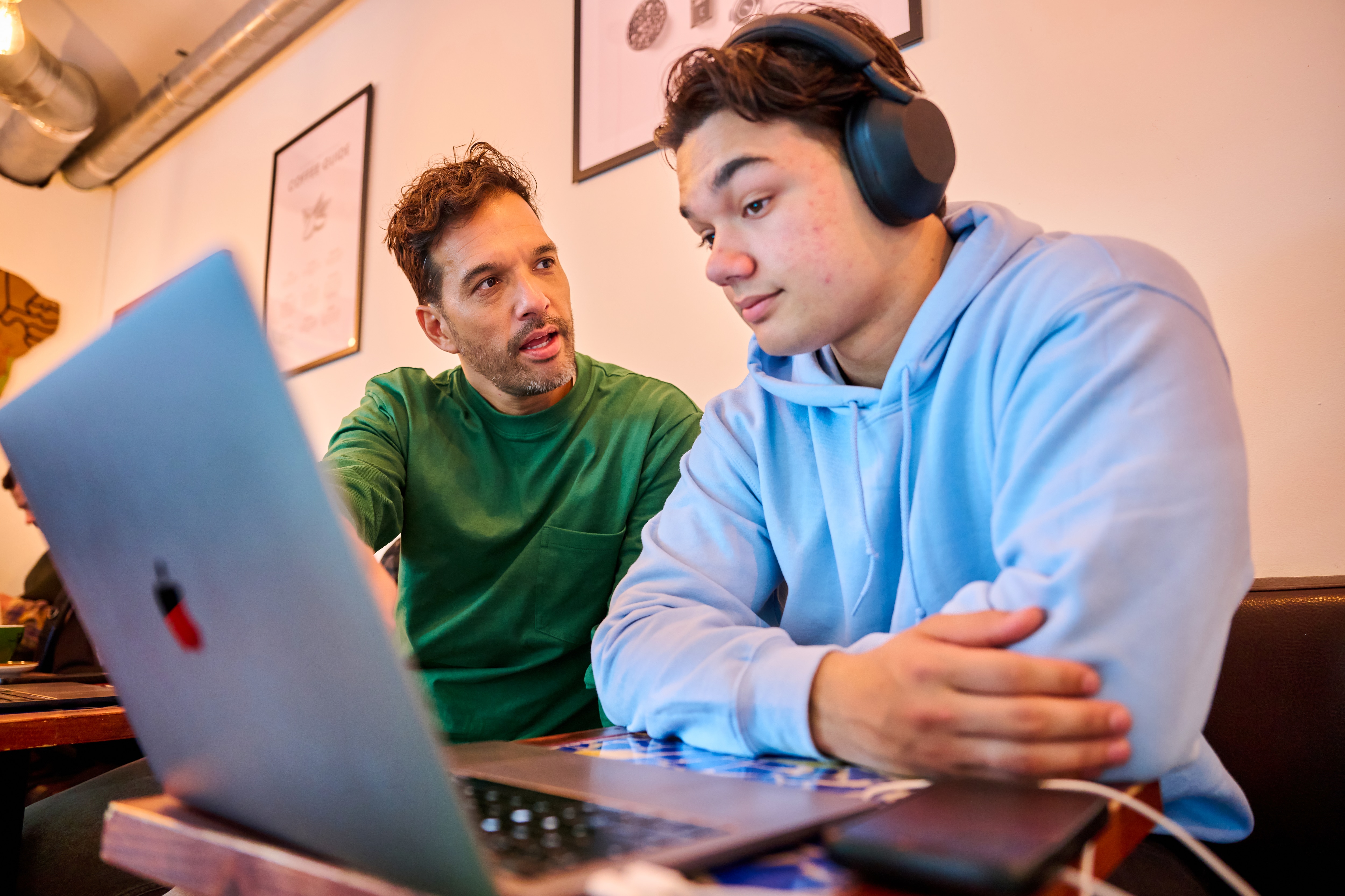 Two men sitting indoors at a small table. One man is wearing headphones and looking at a laptop, while the other points at the screen.