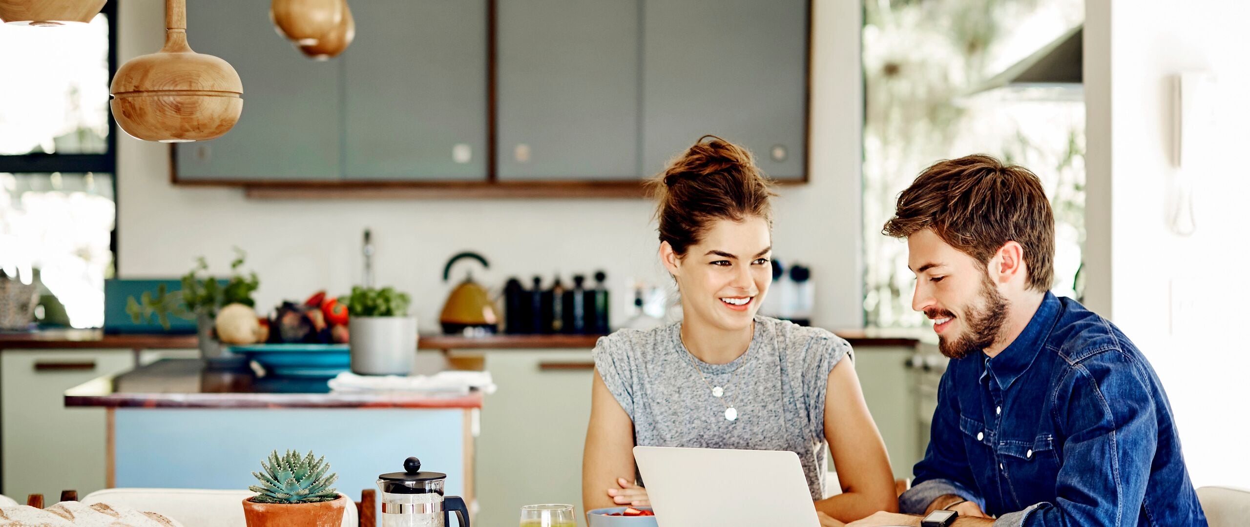 Young male and female bloggers using laptop. Happy couple wearing trendy casuals at home. They are sitting at table with kitchen in background.