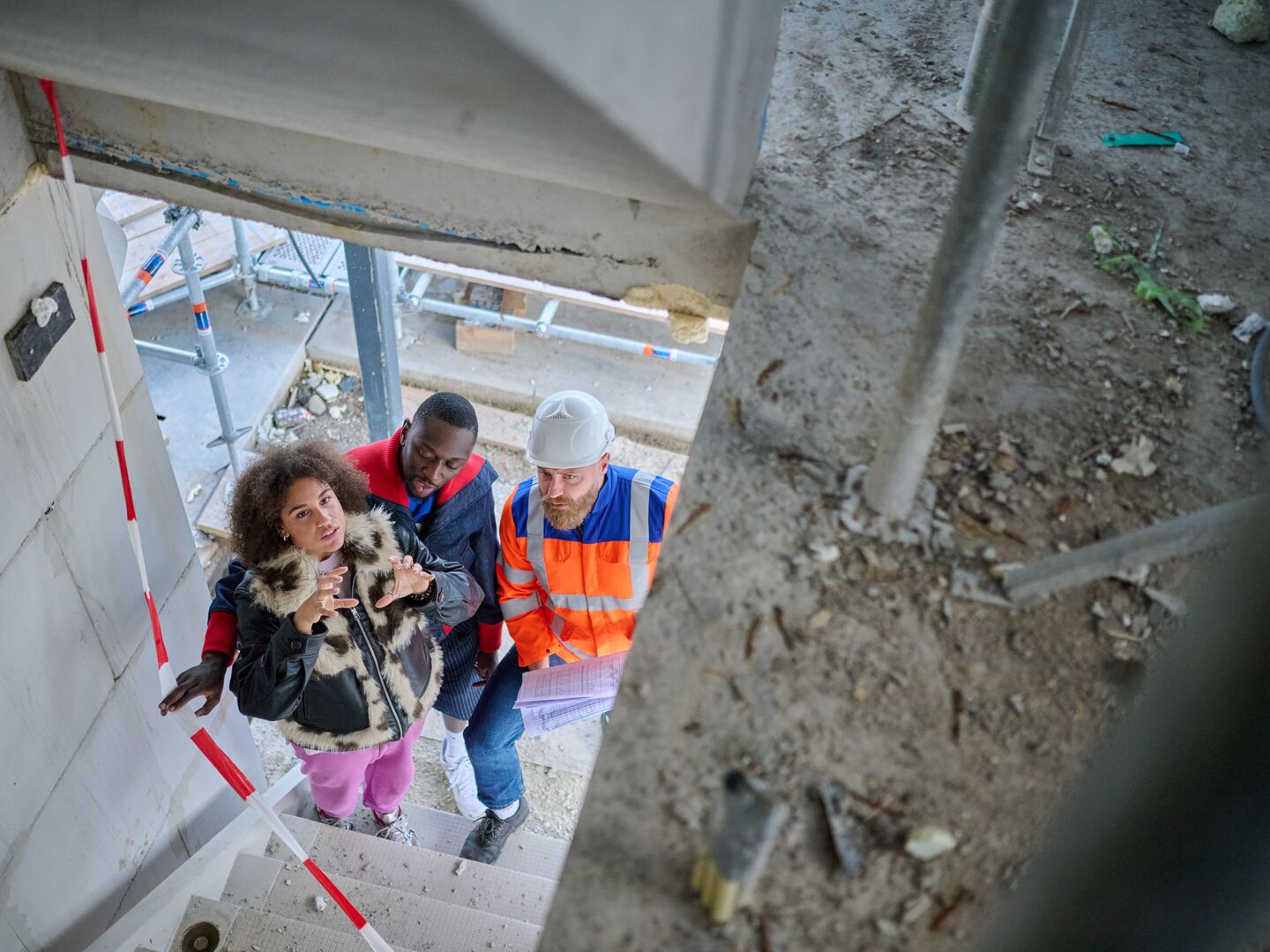 Man and woman inspect the interior of a house under construction with metal support poles and unfinished walls.