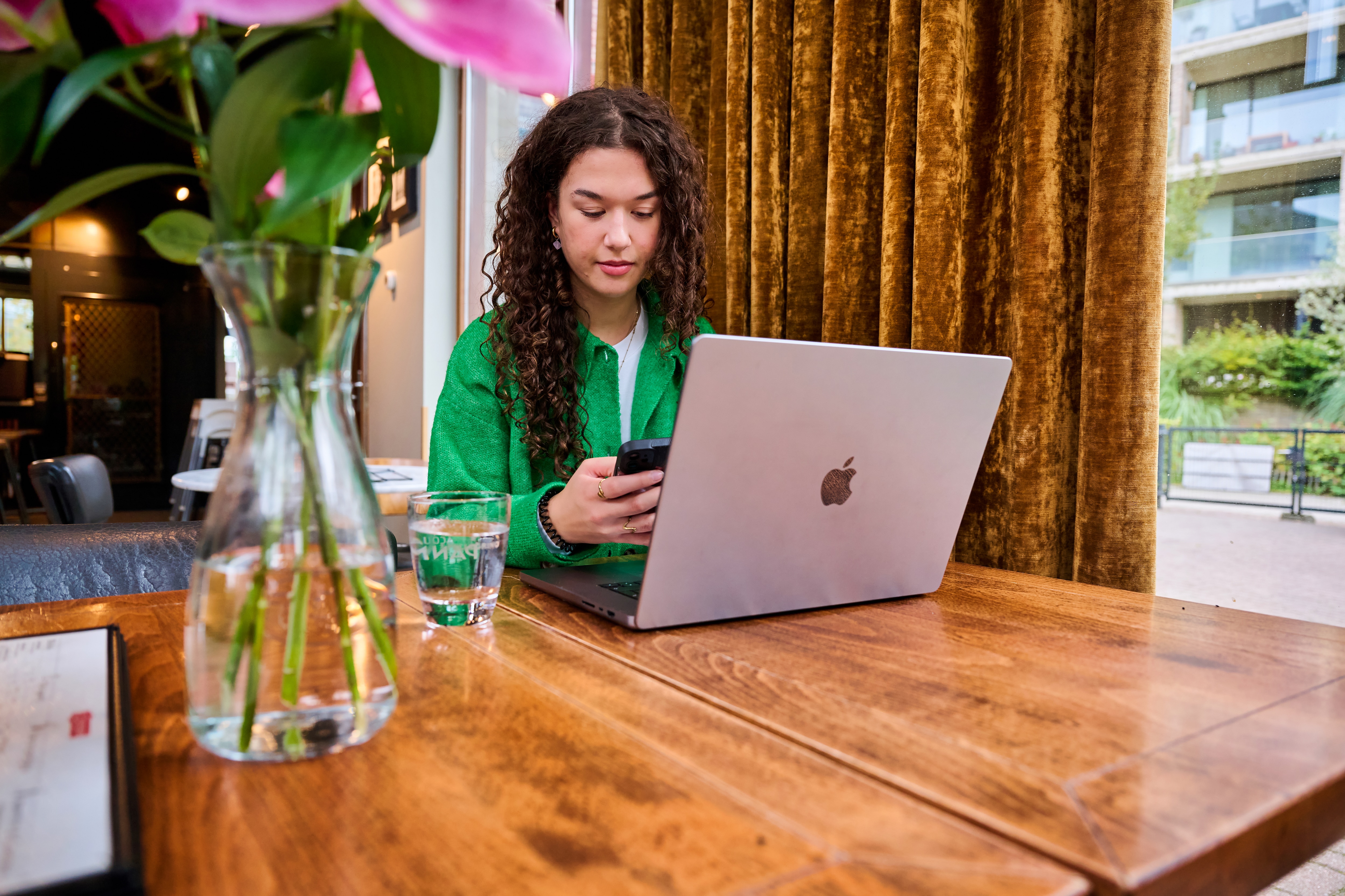 Woman in a green blouse sitting at a table behind a laptop, looking at her phone.