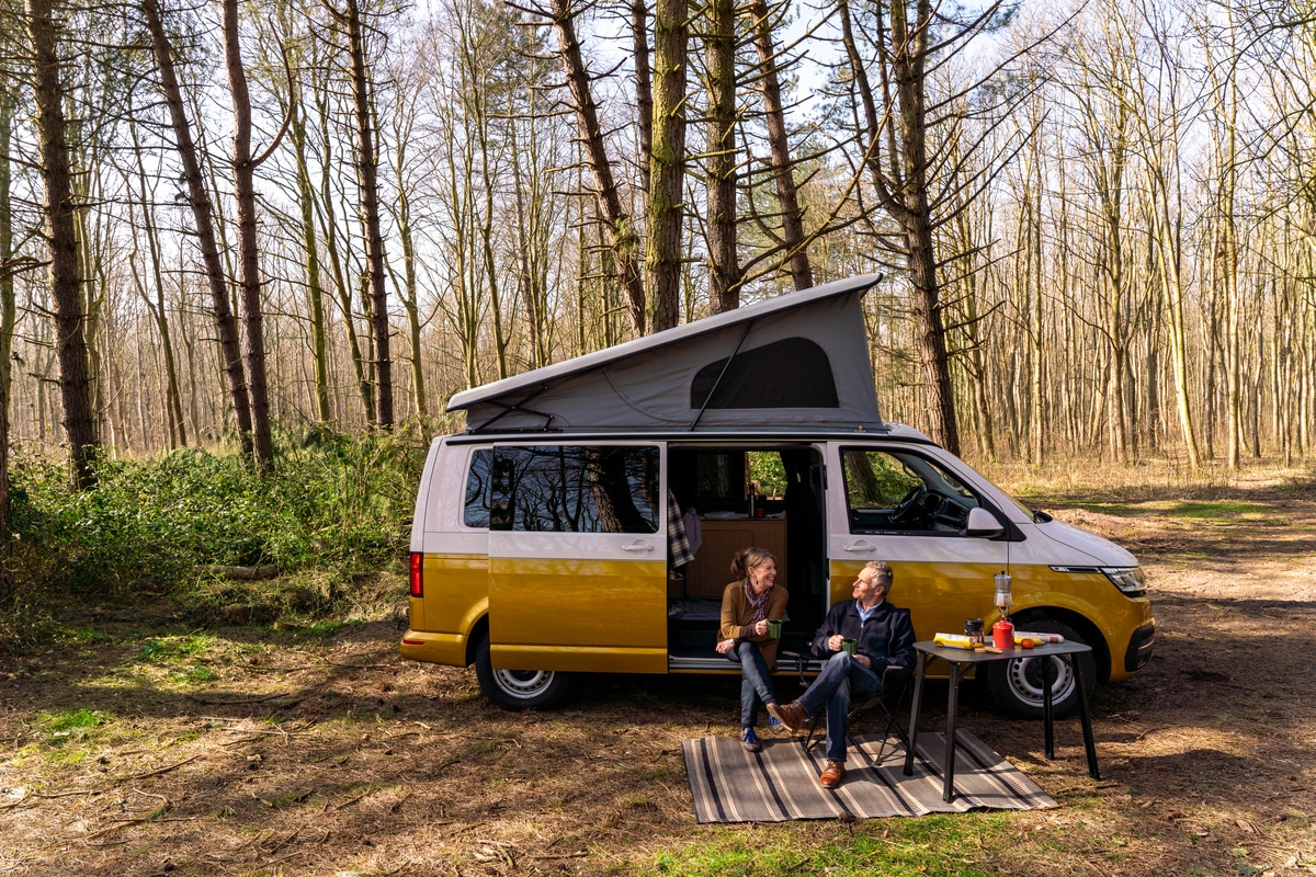 2 mensen die voor een camper zitten in de natuur. Ze drinken koffie en zijn aan het kamperen.